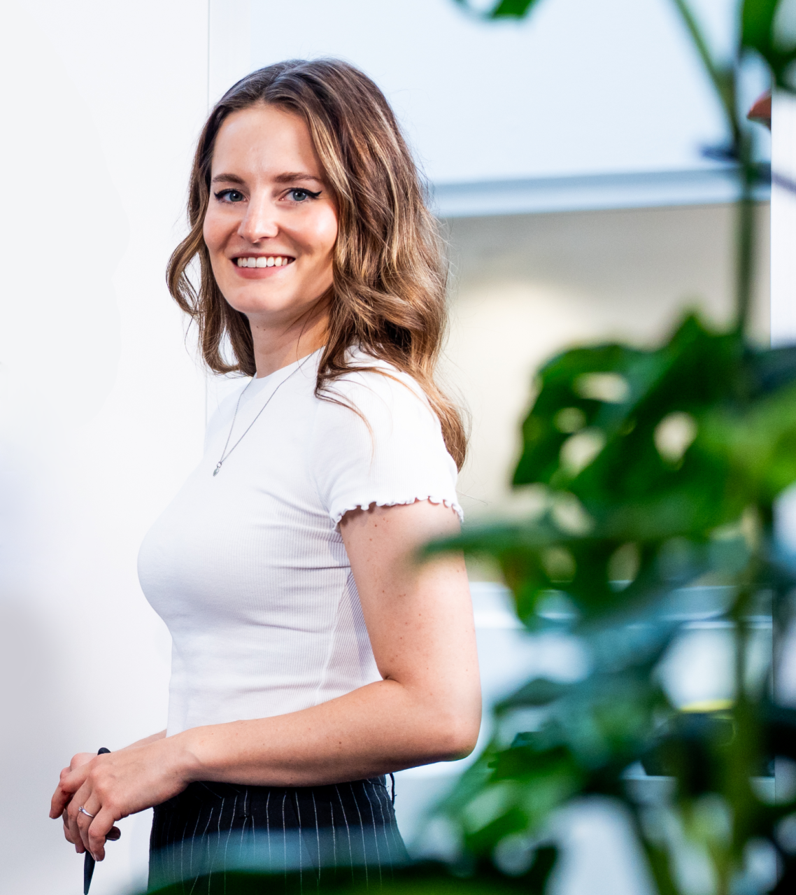 Lächelnde Frau mit langen Haaren, trägt ein weißes Oberteil und steht vor einer Pflanze.