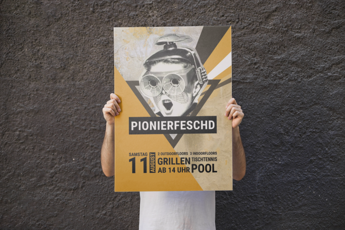Person hält ein Plakat mit der Aufschrift „Pionierfeschd" und weiteren Informationen. Das Plakat dient als Einladung zu einer Feier.